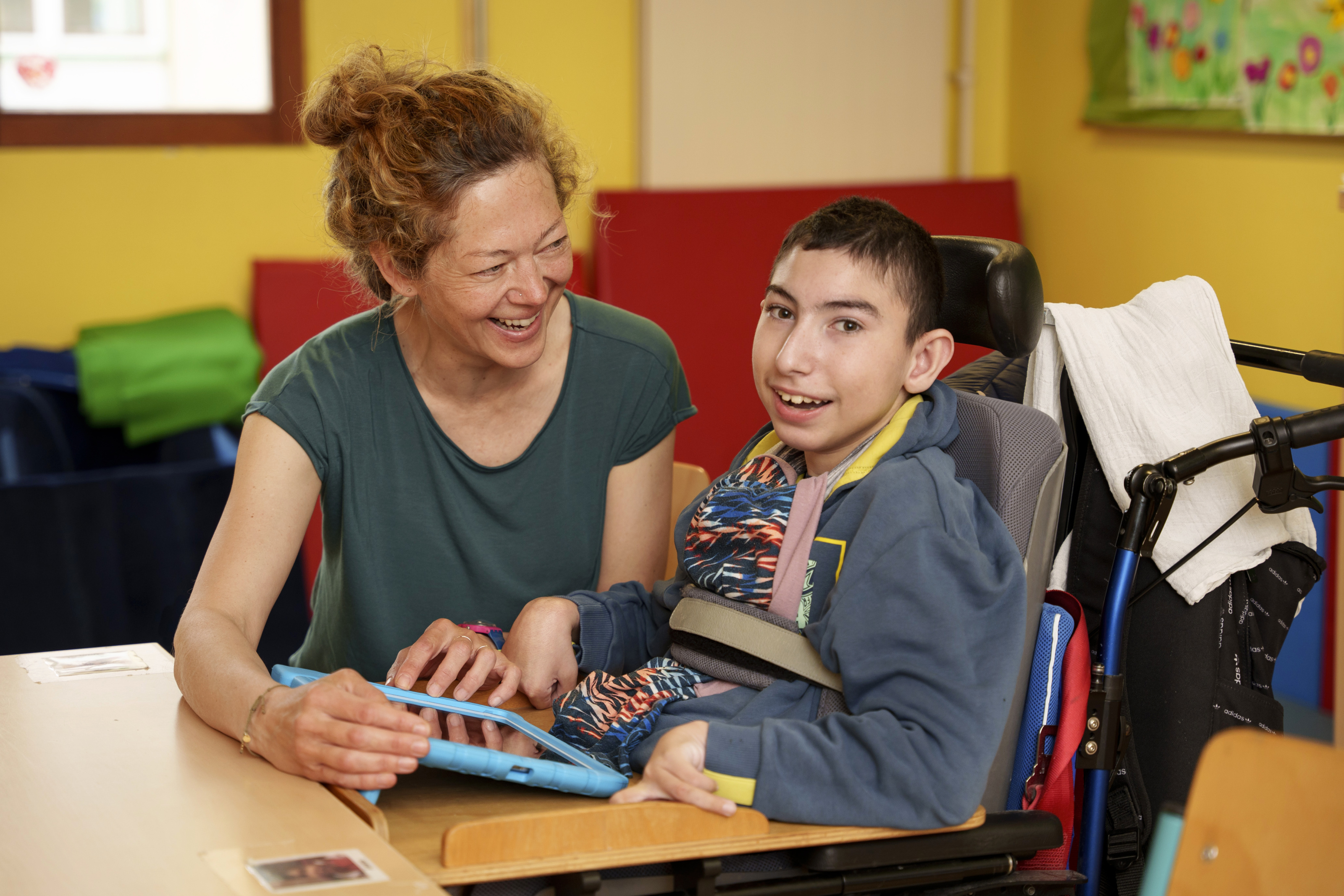 Ein Schüler im Rollstuhl sitzt lächelnd mit einer Betreuungsperson an einem Tisch und arbeitet an einem Tablet.